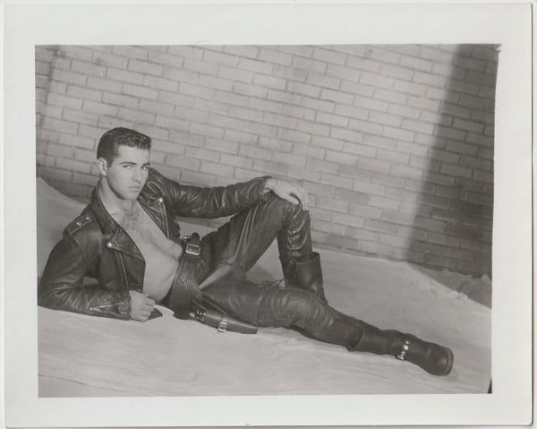 A black and white vintage gay photograph featuring a man in a leather jacket and leather pants reclining in front of a wall.