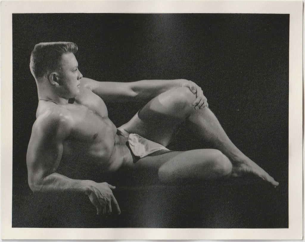 A black and white vintage physique photograph featuring a nude muscular male model reclining against a dark studio background attributed to Bruce of LA.