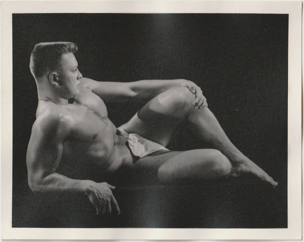 A black and white vintage physique photograph featuring a nude muscular male model reclining against a dark studio background attributed to Bruce of LA.