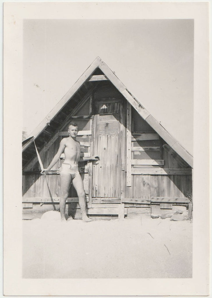 A vintage gay black and white photograph showing a young man in a swimsuit standing barefoot in front of a rustic wooden A-frame cabin.