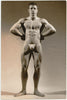 Muscular man with arms akimbo poses for a studio shot by Royale. Vintage gay physique photo.