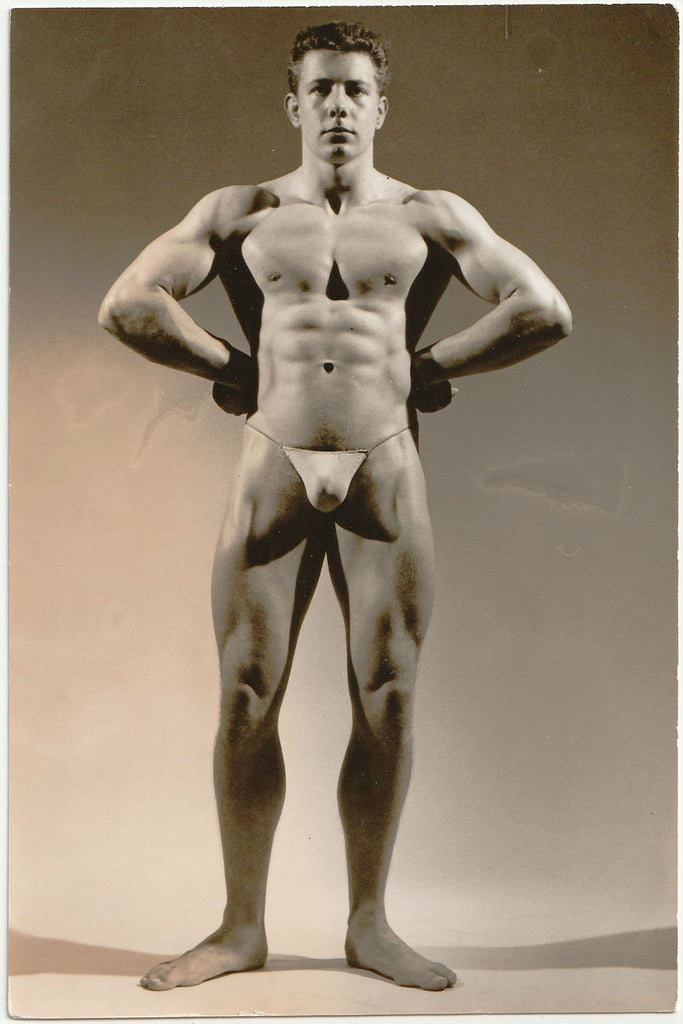 Muscular man with arms akimbo poses for a studio shot by Royale. Vintage gay physique photo.