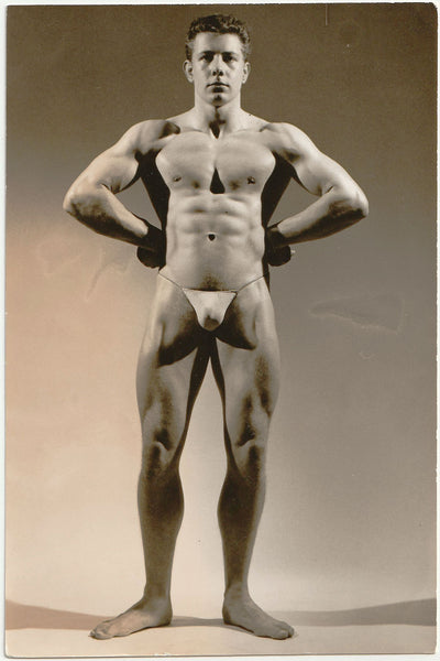 Muscular man with arms akimbo poses for a studio shot by Royale. Vintage gay physique photo.
