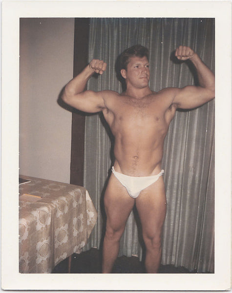 Vintage gay Polaroid Handsome, muscular man flexes his biceps while he stands next to the dining room table vintage gay color Polaroid