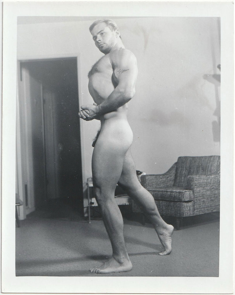 Barely contained in the frame of the photo, a muscular guy with "Jim" tattooed on his arm stands naked in the living room.
