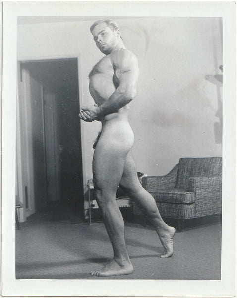 Barely contained in the frame of the photo, a muscular guy with "Jim" tattooed on his arm stands naked in the living room.