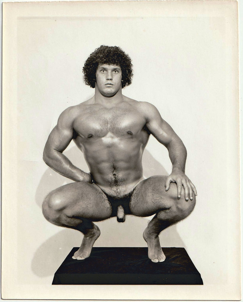 Big boy with curly hair stares into the camera as he crouches on a plinth. Vintage gay physique photo.