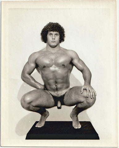 Big boy with curly hair stares into the camera as he crouches on a plinth. Vintage gay physique photo.