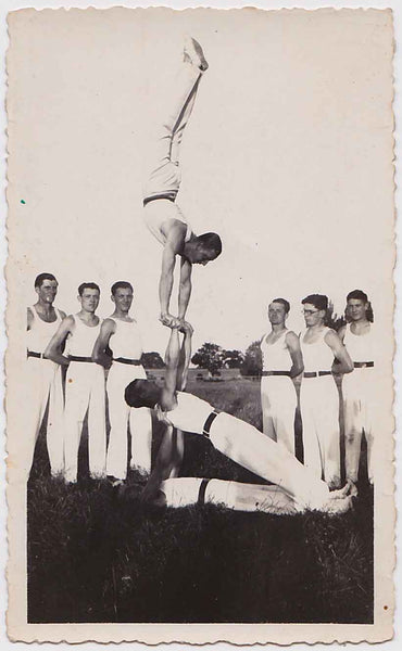 A team of nine gymnasts. Six stand with arms behind their backs watching three others perform. Vintage photo.