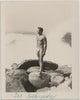 A vintage black and white photograph featuring a nude man, Joe Schneider, standing on large rocks by the ocean.
