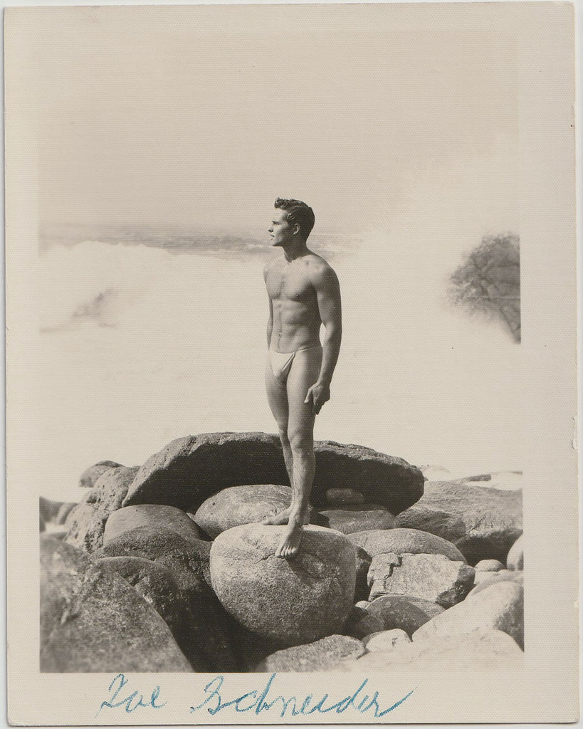 A vintage black and white photograph featuring a nude man, Joe Schneider, standing on large rocks by the ocean.