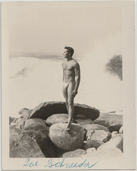 A vintage black and white photograph featuring a nude man, Joe Schneider, standing on large rocks by the ocean.