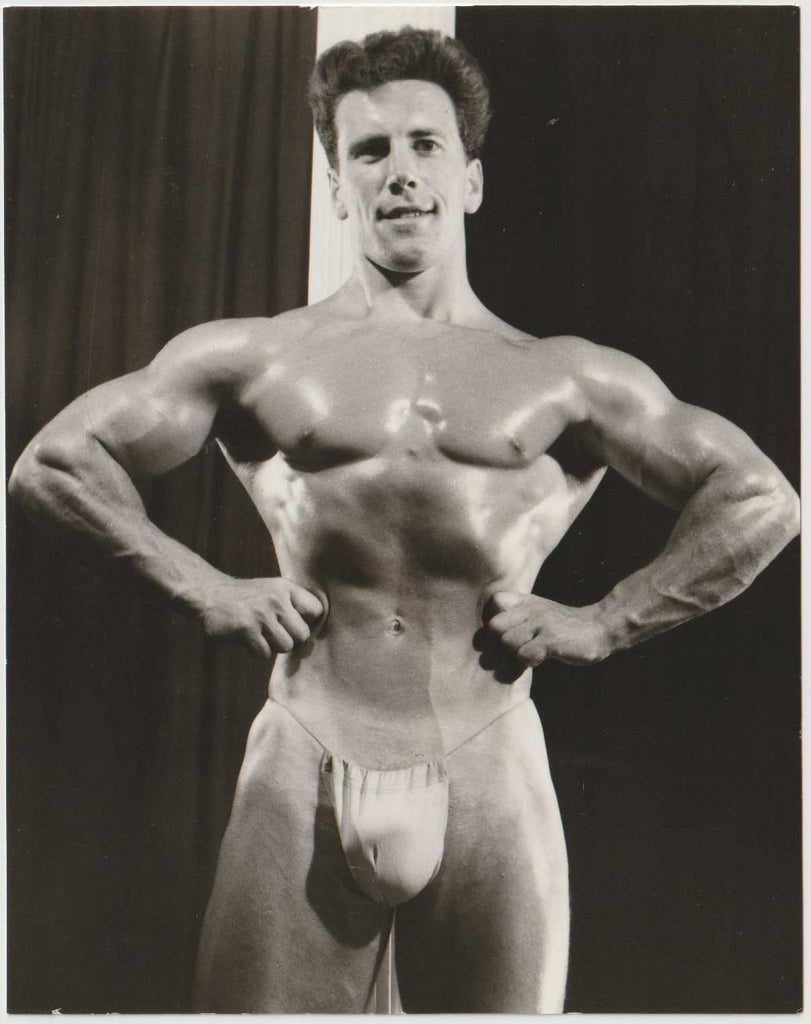 Lightly oiled and standing in front of a column, the model in posing strap with arms akimbo Johnny Francis.