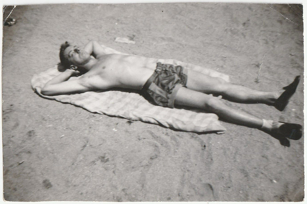 Black and white vintage photograph of a man lying on a towel on a sandy beach, wearing patterned shorts and dark loafers.