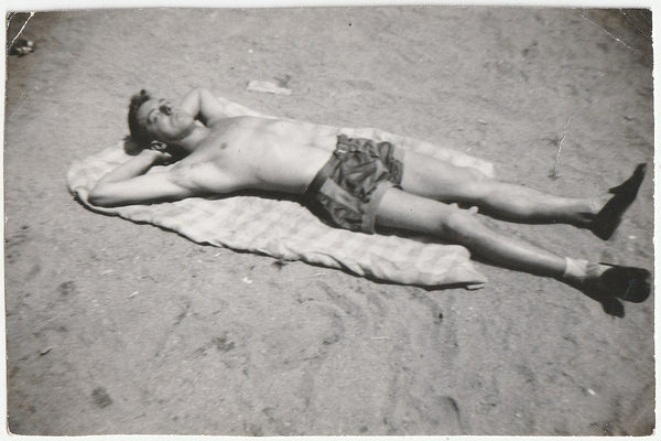 Black and white vintage photograph of a man lying on a towel on a sandy beach, wearing patterned shorts and dark loafers.