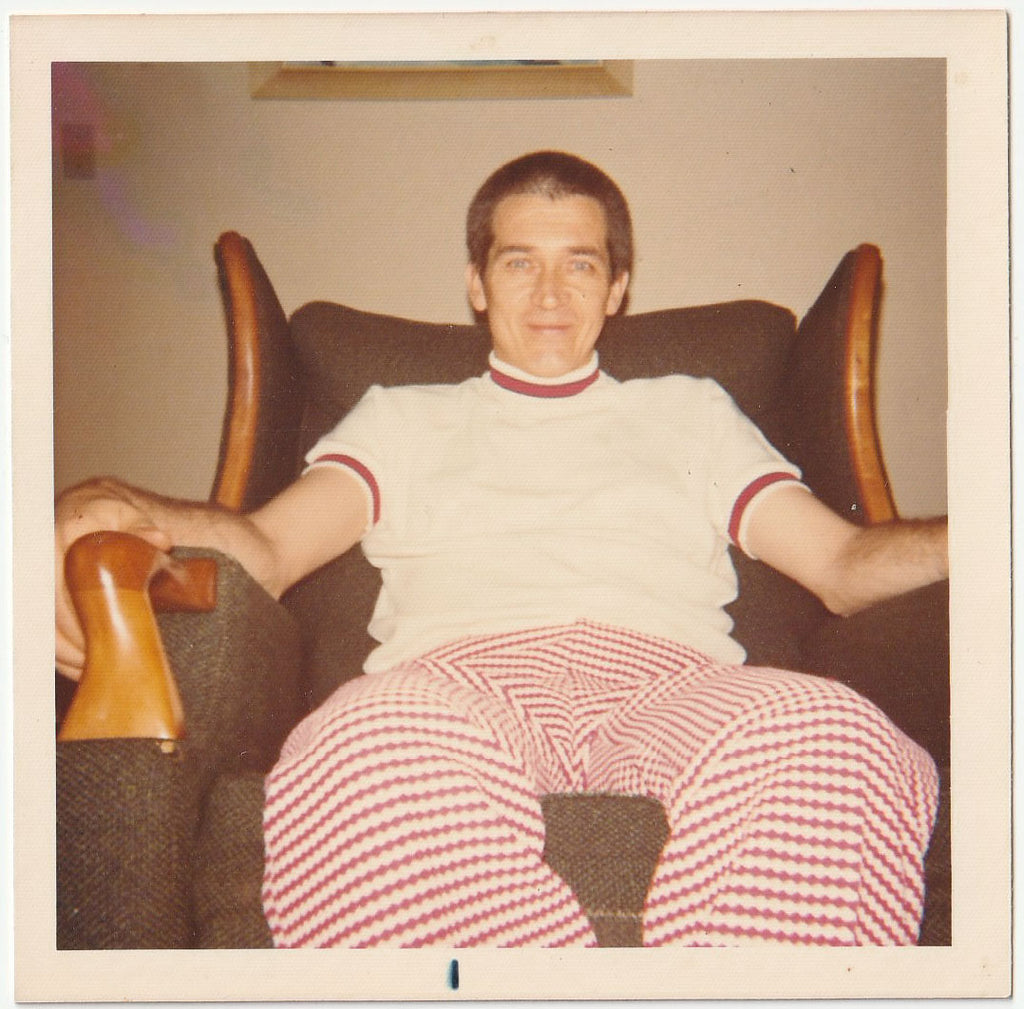 A vintage-style color photograph of a man sitting in a dark armchair, wearing a white short-sleeve turtleneck sweater with red trim and red-and-white checkered pants.