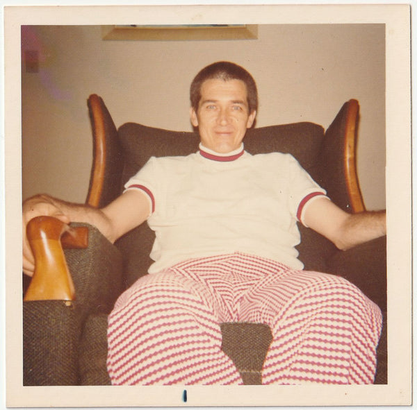 A vintage-style color photograph of a man sitting in a dark armchair, wearing a white short-sleeve turtleneck sweater with red trim and red-and-white checkered pants.