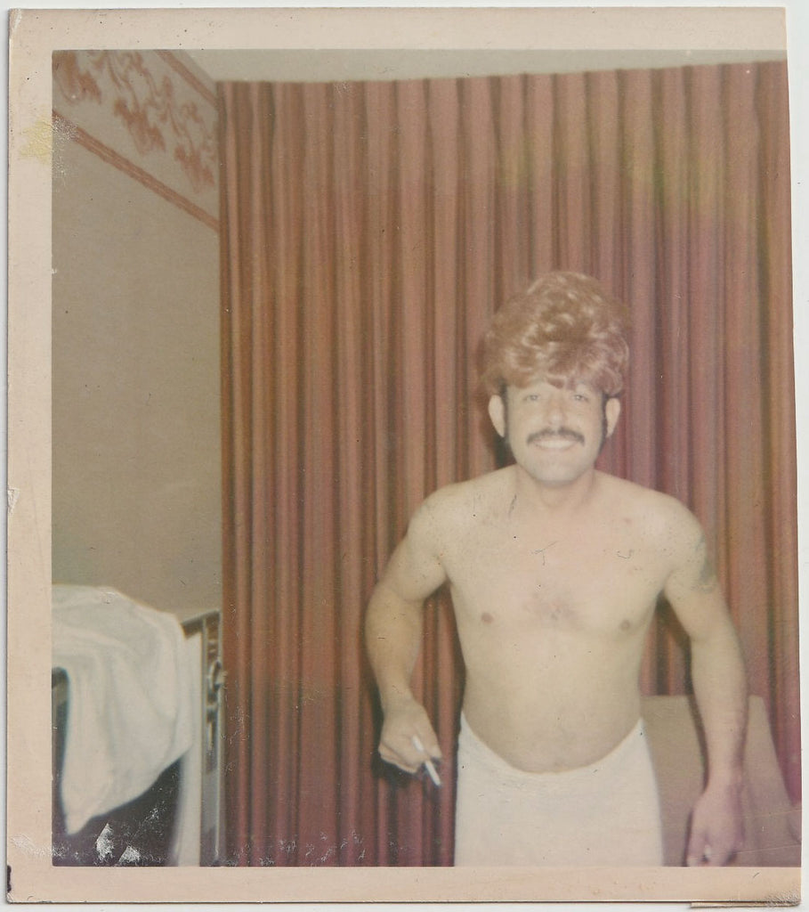 Cute mustachioed guy wearing a towel and a blond wig in what looks like a motel room vintage color snapshot.