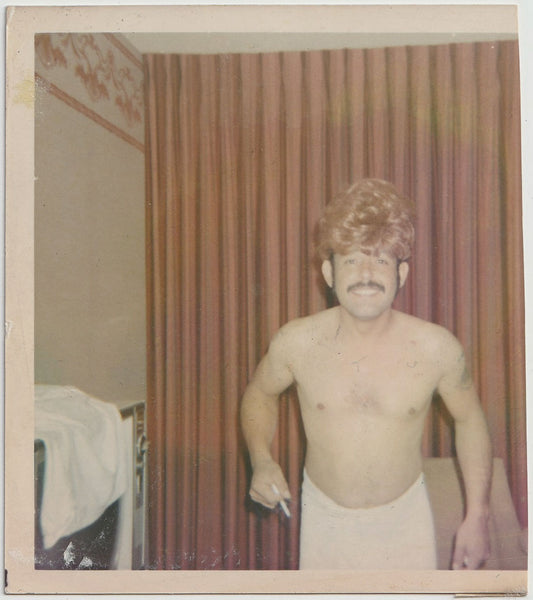 Cute mustachioed guy wearing a towel and a blond wig in what looks like a motel room vintage color snapshot.