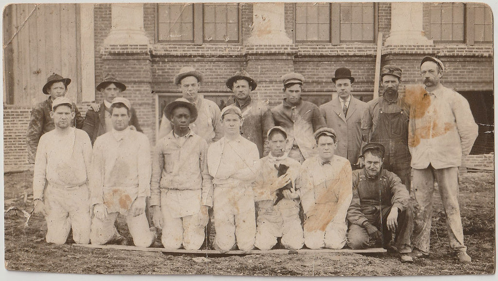 Real Photo Postcard of men in two rows, posing in front of a brick building. C. 1912.