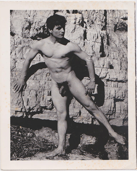 Handsome male nude balances on one leg as he touches the rock face behind him. Vintage gay photo