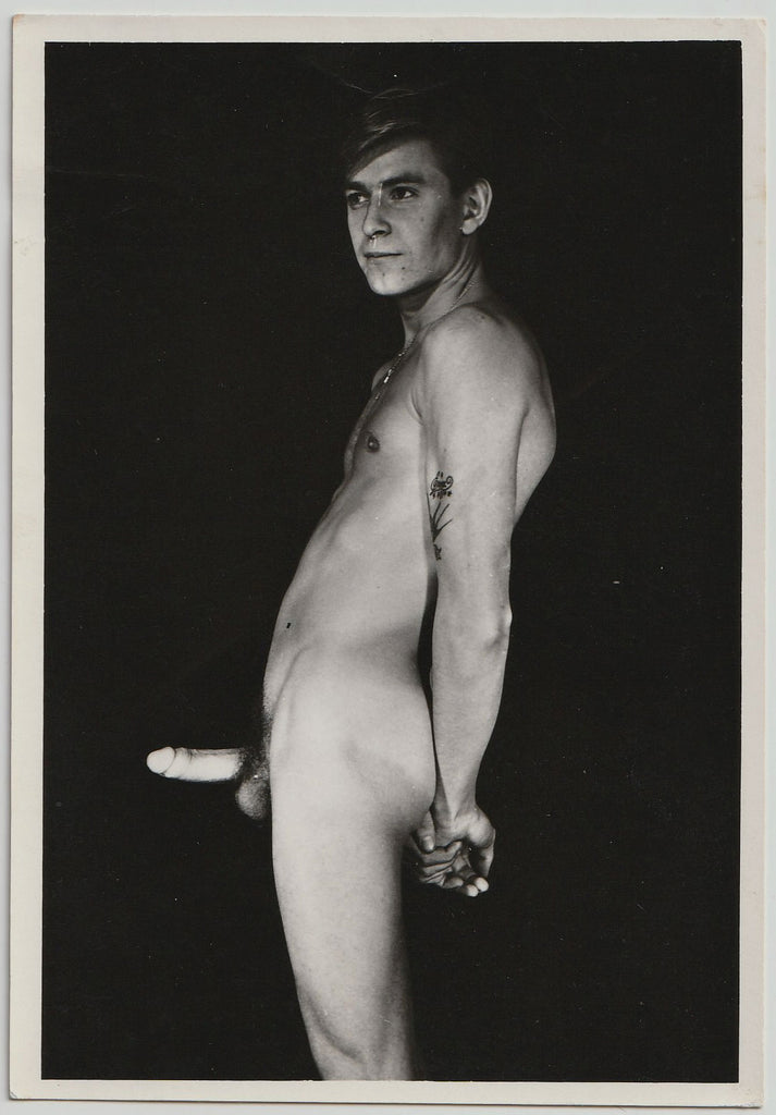 Leaning back, hands tightly clasped, faraway expression, this nude man has a floral tattoo on his arm and what looks like a glow-in-the-dark dick.
