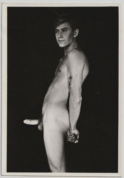 Leaning back, hands tightly clasped, faraway expression, this nude man has a floral tattoo on his arm and what looks like a glow-in-the-dark dick.