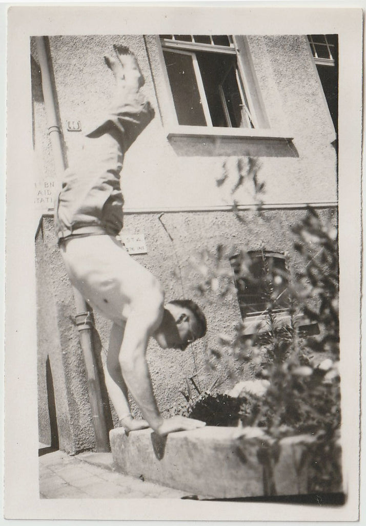A vintage black and white photograph depicting a shirtless and shoeless man performing a handstand on a concrete curb.