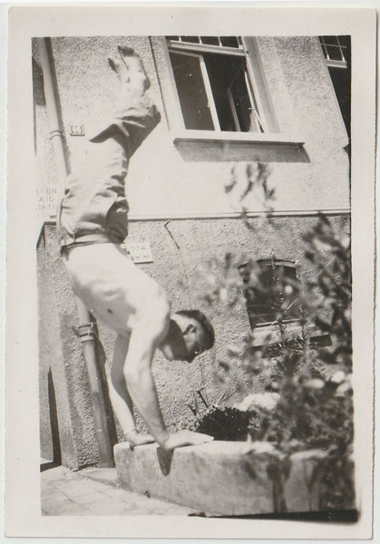A vintage black and white photograph depicting a shirtless and shoeless man performing a handstand on a concrete curb.