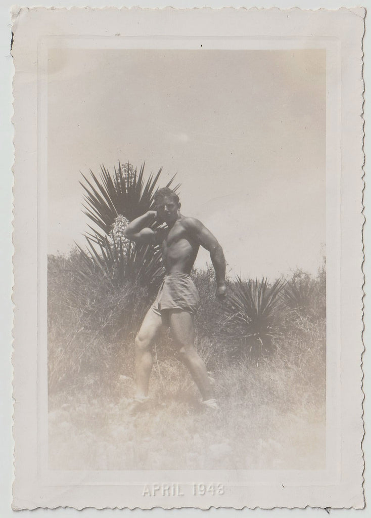 A vintage sepia photograph print showing a shirtless man flexing in front of a flowering yucca.