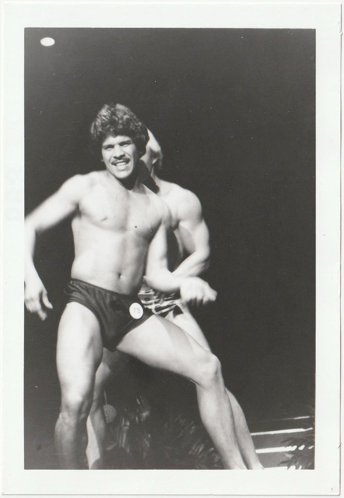 A black and white photograph of a muscular male bodybuilder posing on stage in shorts, with another contestant partially visible behind him.