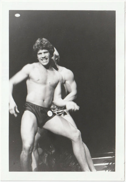 A black and white photograph of a muscular male bodybuilder posing on stage in shorts, with another contestant partially visible behind him.