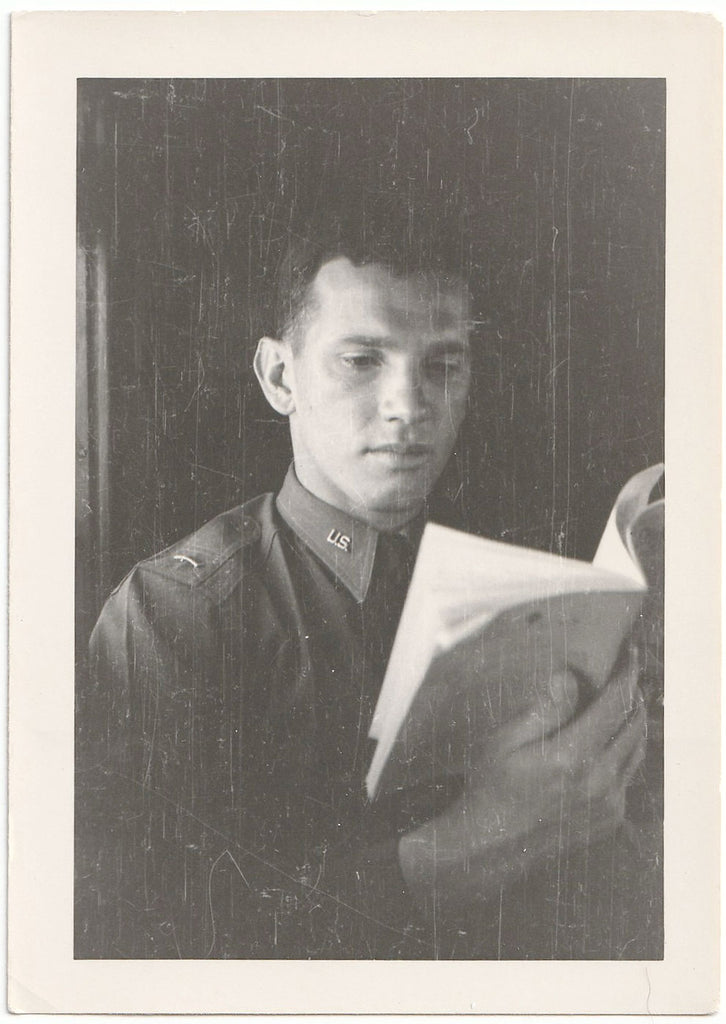 A vintage black and white portrait photograph print of a young man in a military uniform holding an open book.
