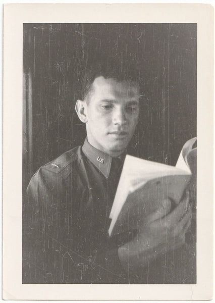 A vintage black and white portrait photograph print of a young man in a military uniform holding an open book.