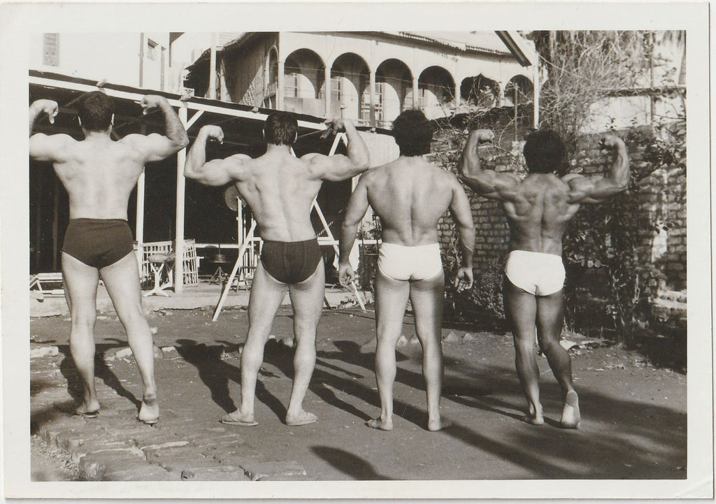 A black and white photograph showing four muscular men from behind, flexing in an outdoor setting with a building and brick wall in the background.