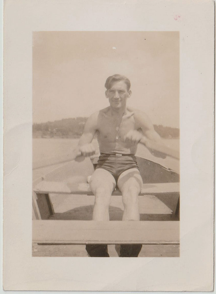 A vintage sepia-toned photograph print showing a handsome man seated in a rowboat wearing dark swim trunks. 