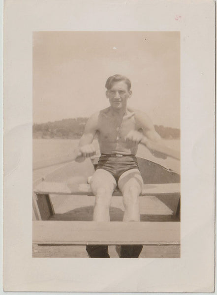 A vintage sepia-toned photograph print showing a handsome man seated in a rowboat wearing dark swim trunks. 