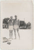 A vintage gay black and white photograph showing two affectionate men standing on a beach, wearing swim trunks. 