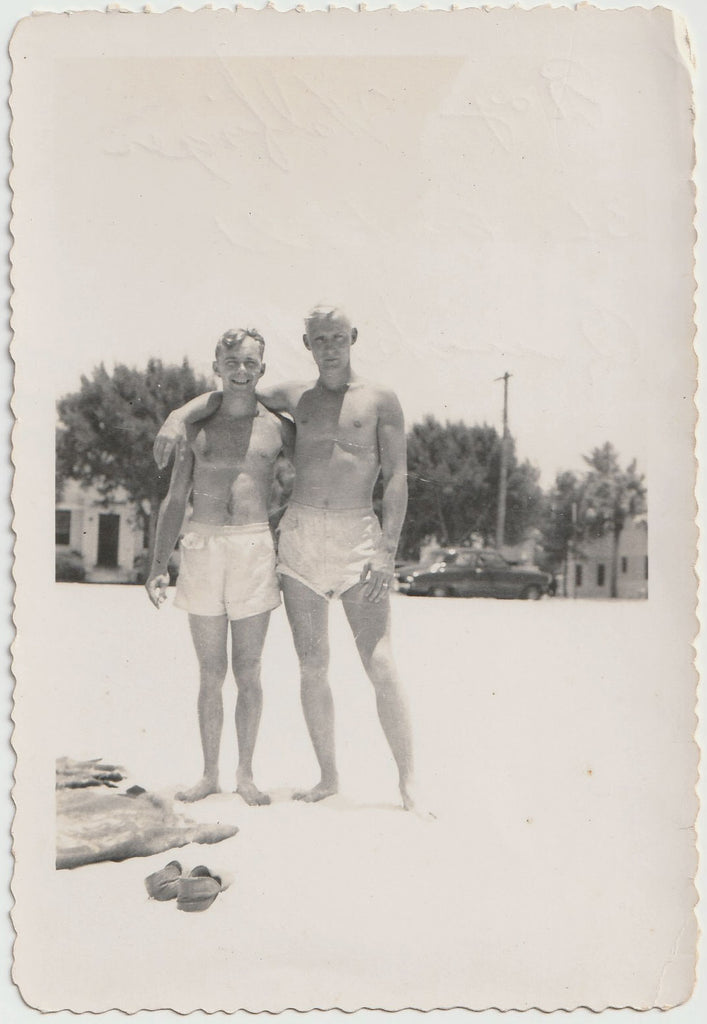 A vintage gay black and white photograph showing two affectionate men standing on a beach, wearing swim trunks. 
