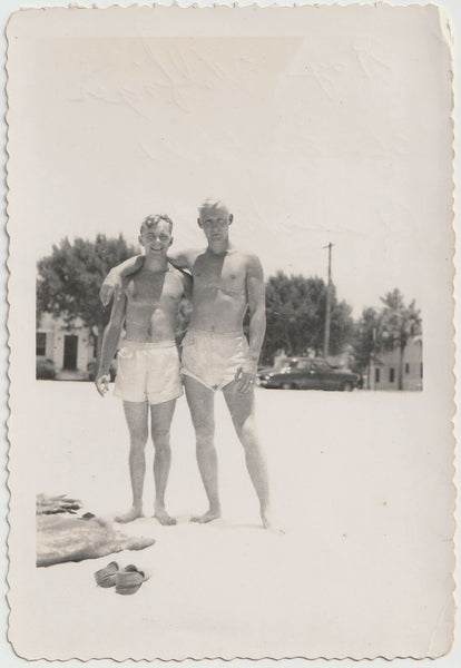 A vintage gay black and white photograph showing two affectionate men standing on a beach, wearing swim trunks. 
