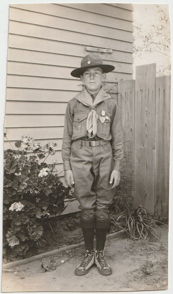 Vintage black and white photo of a sad boy in scout uniform standing next to his house.