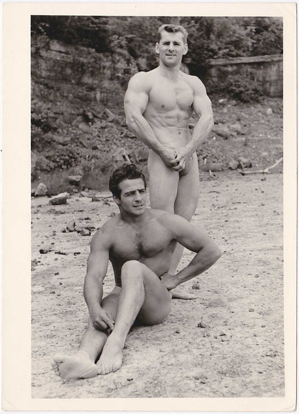 Bodybuilder duo. Handsome guys, one seated and relaxed, one standing and flexing on the sand. Vintage gay photo.