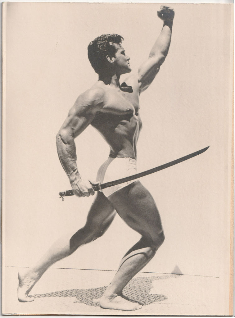 Vintage photo by an unidentified photographer shows Steve Reeves raising a fist and holding a sword as he lunges forward.
