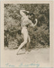 A vintage black and white photograph print showing a muscular nude man, Tim Sweeny, posing outdoors against a background of dense foliage. Physique photo c. 1958 by Spectrum.