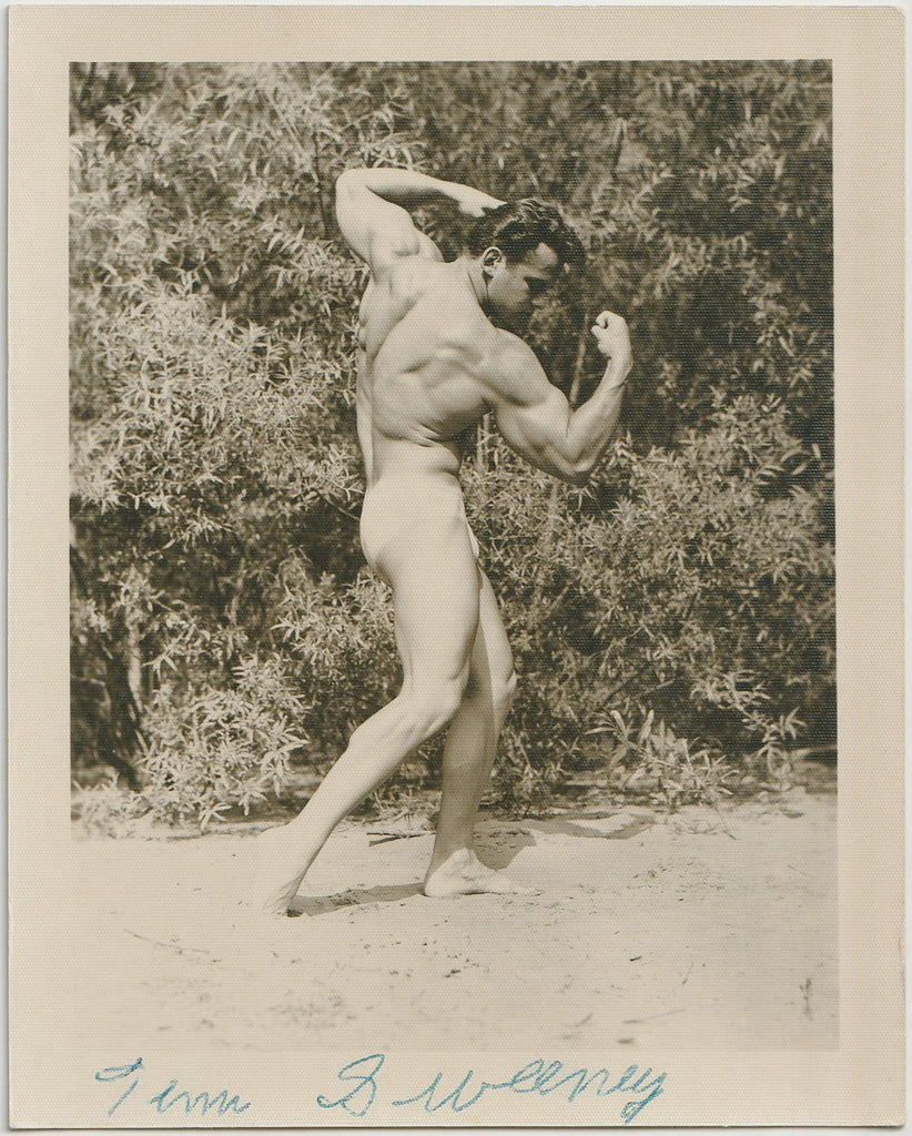 A vintage black and white photograph print showing a muscular nude man, Tim Sweeny, posing outdoors against a background of dense foliage. Physique photo c. 1958 by Spectrum.