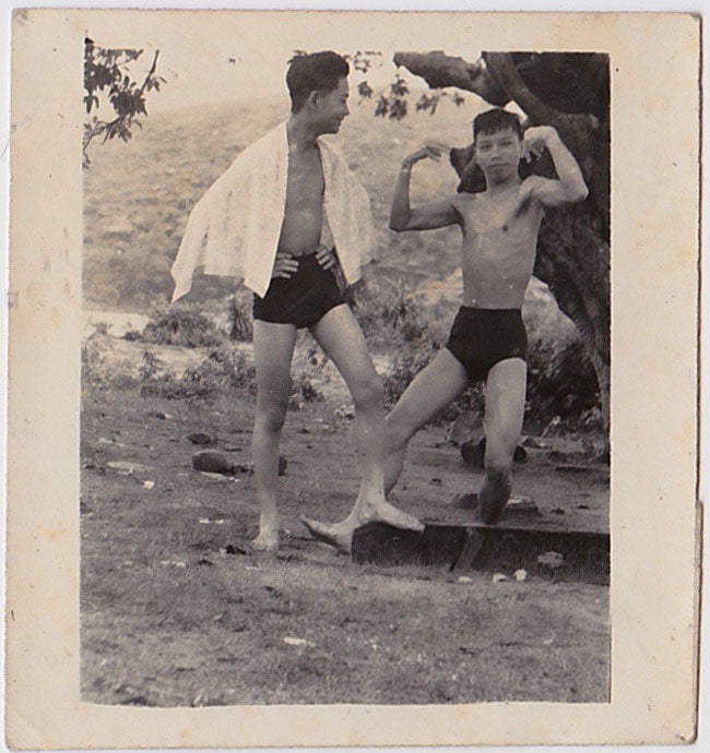 Undated vintage photo of two Asian men in swimsuits, one of whom watches the other flexing his biceps.