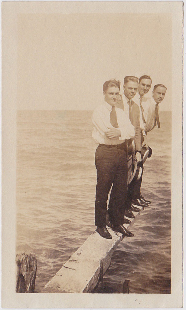 Four cute young guys wearing shirts and ties, carrying straw boaters, stand suspended over the water vintage photo