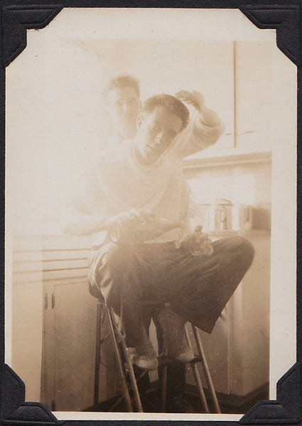 Two men in the kitchen. One cuts the other's hair while he's pouring something from a bottle into his glass.