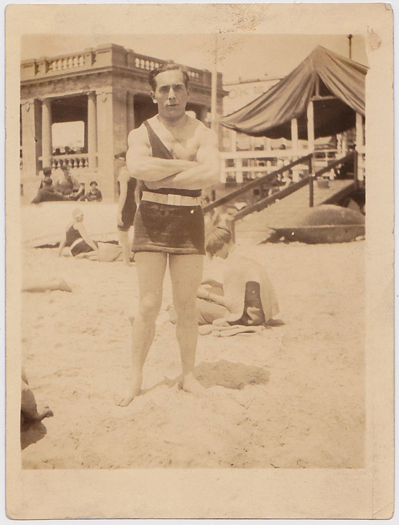 Undated vintage sepia photo of a bodybuilder on the beach in a tough guy pose. 