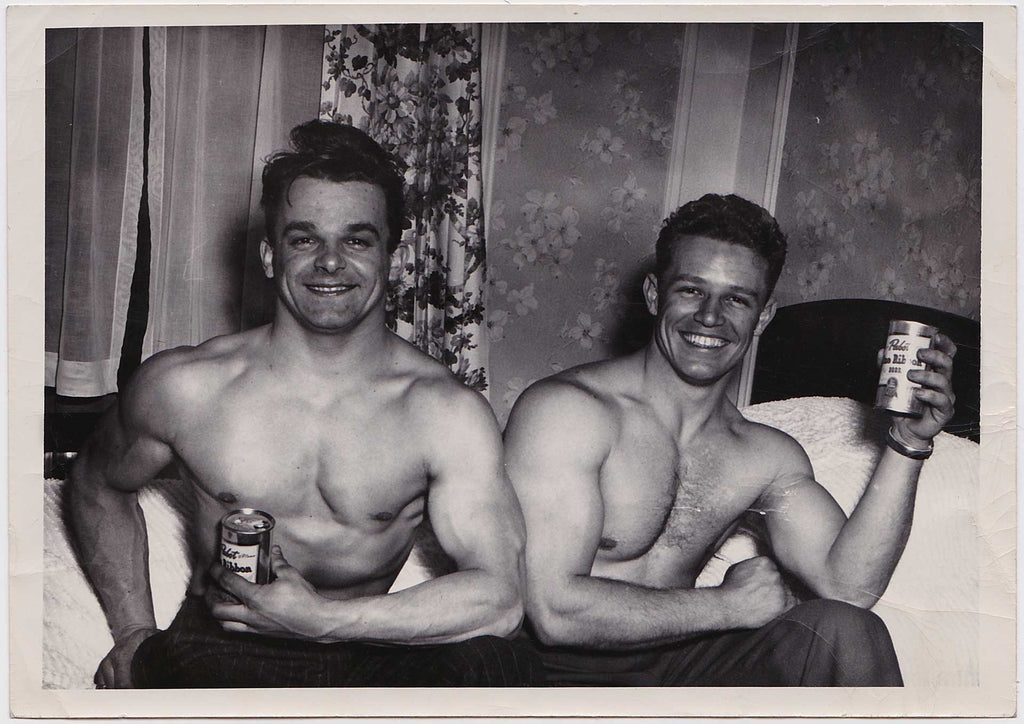 Two good looking, shirtless bodybuilders sit together on the edge of a bed vintage photo